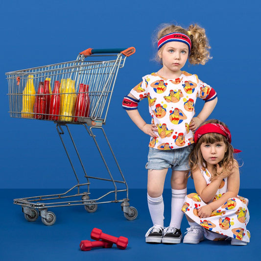 Two young girls modeling matching white t-shirt and dress sets with a colorful cartoon chicken print, posed next to a miniature shopping cart filled with mustard and ketchup bottles on a solid blue studio background.