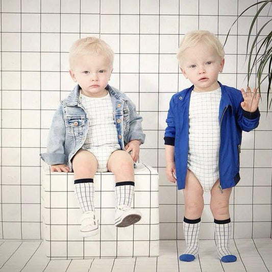 Two toddlers posing in matching white and gray checkered bodysuits with denim jackets. One toddler is sitting on a white tiled block, wearing a light-wash denim jacket, while the other stands next to it wearing a royal blue denim jacket.